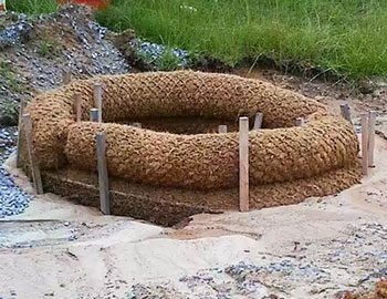 A circular installation of coir logs secured with wooden stakes on sandy soil for erosion control.